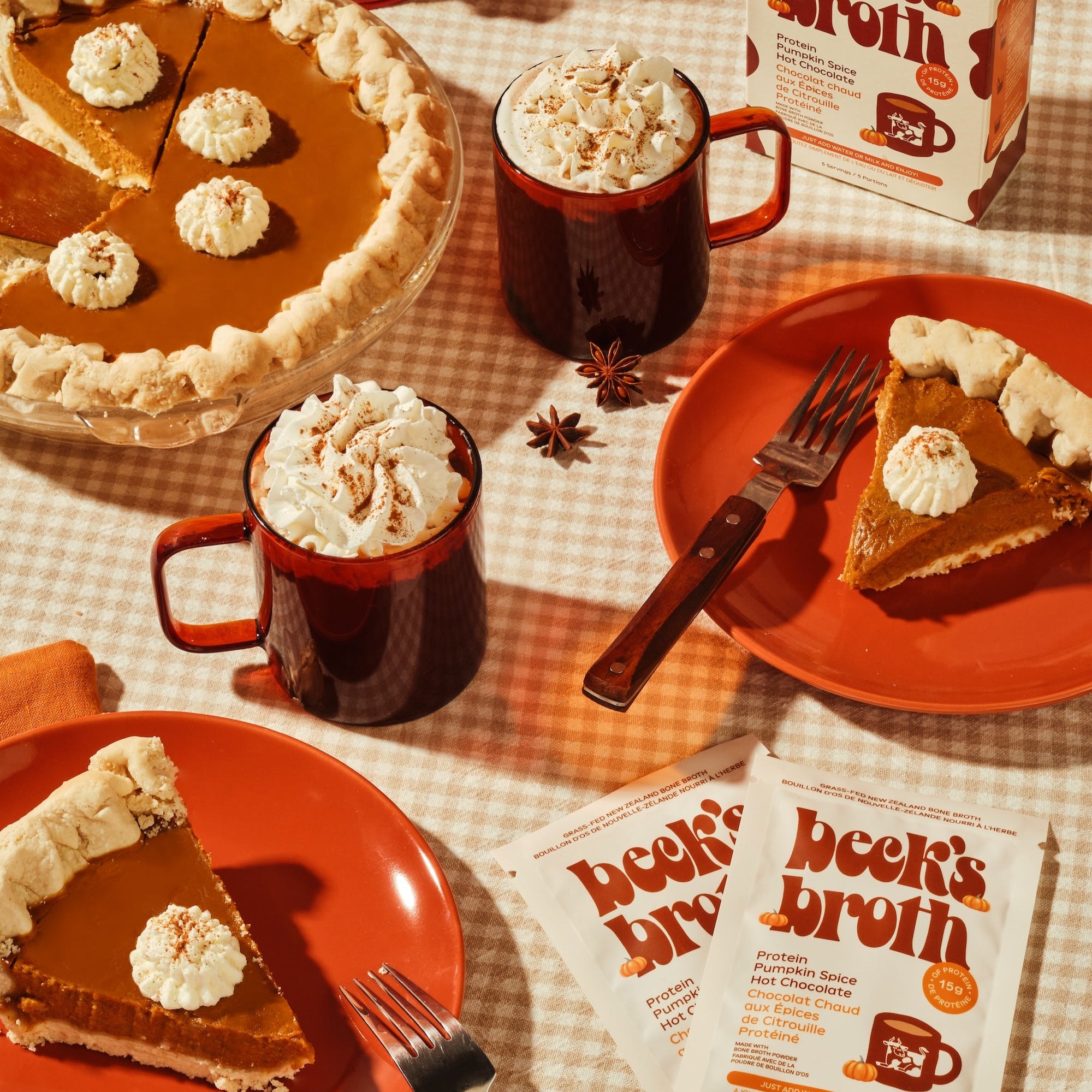 Pumpkin pie with slices on plates, mugs of hot chocolate, and boxes of 'Beck's Broth' on a checkered tablecloth.