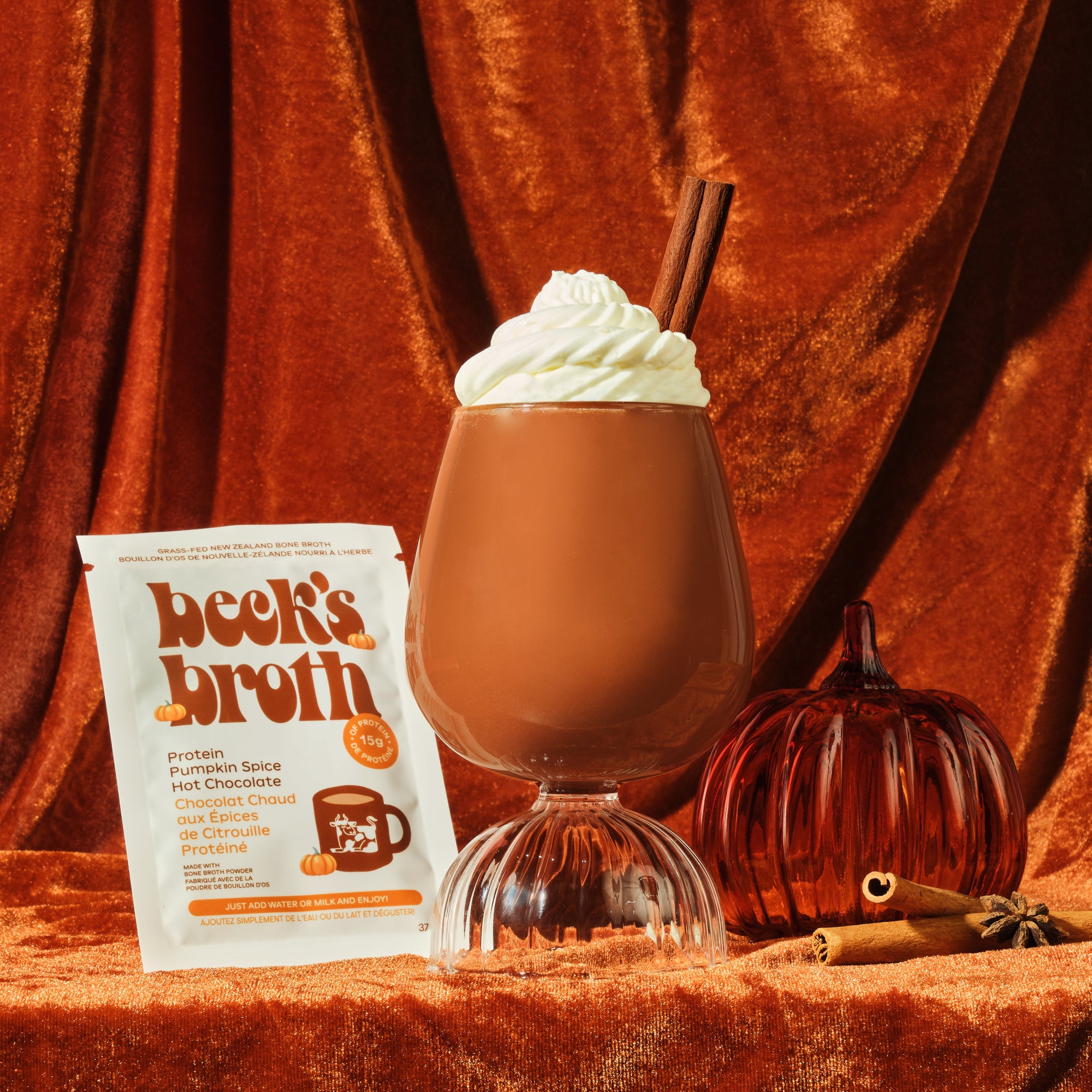 Hot chocolate with whipped cream and a cinnamon stick, accompanied by a package of 'beck's broth' on a rust-colored fabric background.